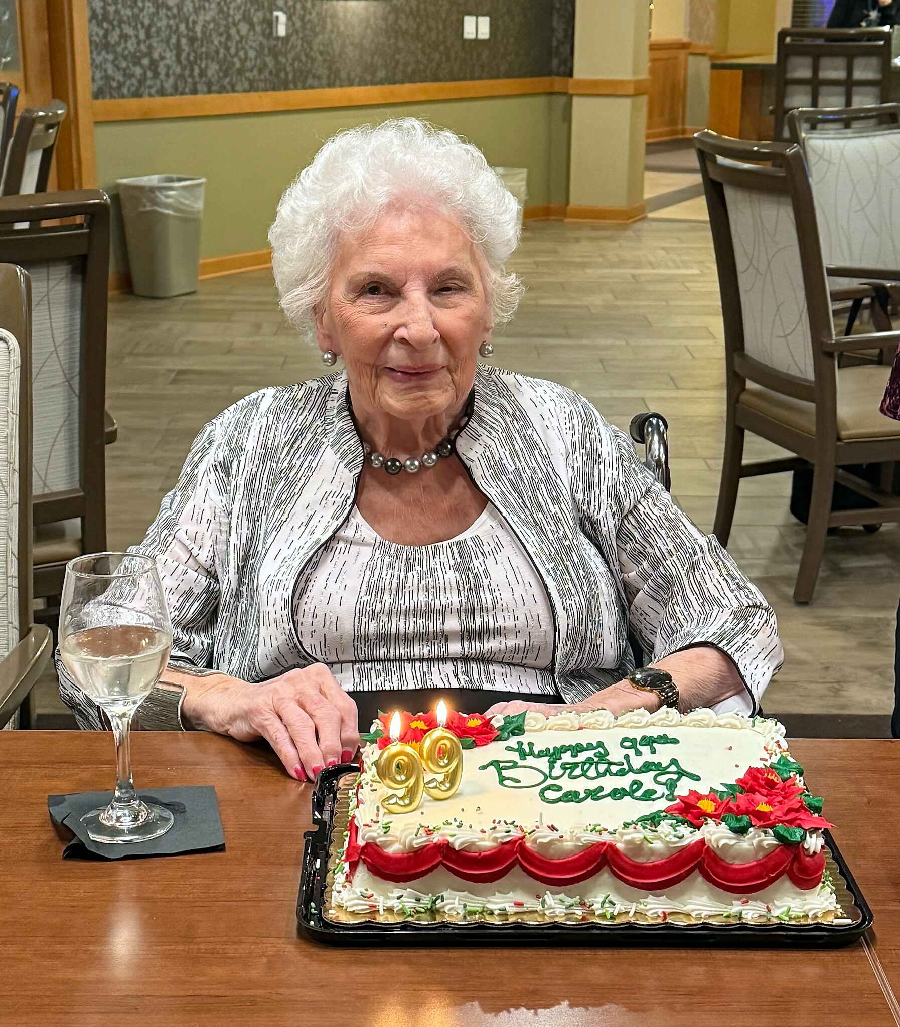 Smiling elderly woman in a striped outfit at a dining table with a decorated birthday cake and candles in front of her.