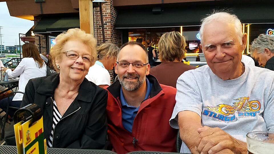 Three people smiling at an outdoor restaurant table: an older woman on the left, a middle-aged man with glasses in the middle, and an older man on the right, with other diners and a brick building in the background.