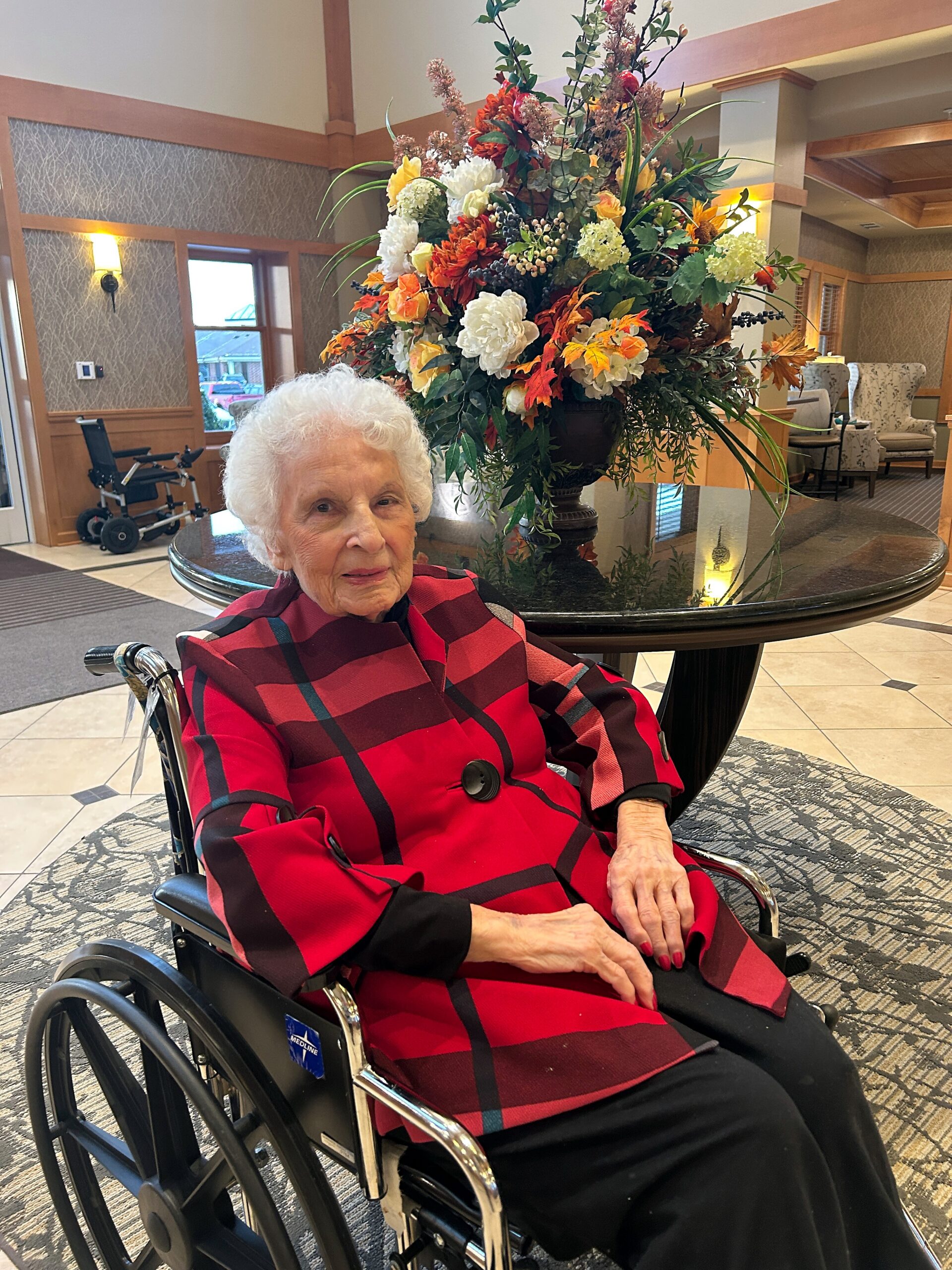 Elderly woman in a red and black plaid jacket sits in a wheelchair beside a large colorful flower arrangement on a round table.