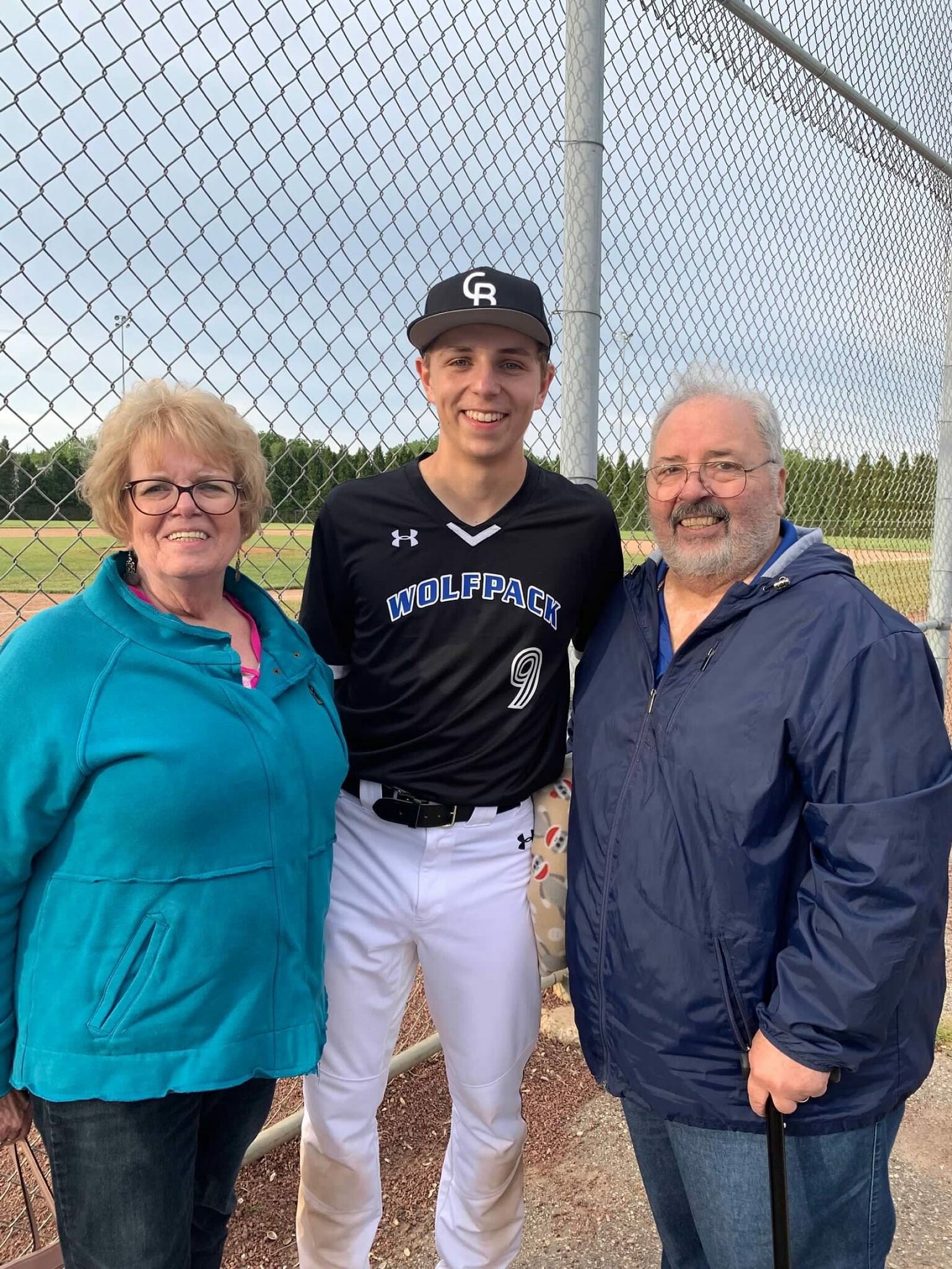 Three people posing at a baseball field, with a chain-link fence behind them; the center person wears a Wolfpack baseball uniform and cap.