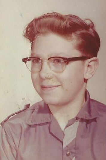 Young person with short wavy hair and glasses, wearing a collared shirt, posing for a vintage portrait.