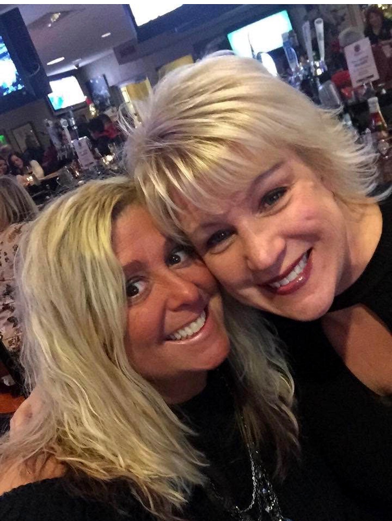 Two smiling women with blond hair taking a close selfie in a busy bar.