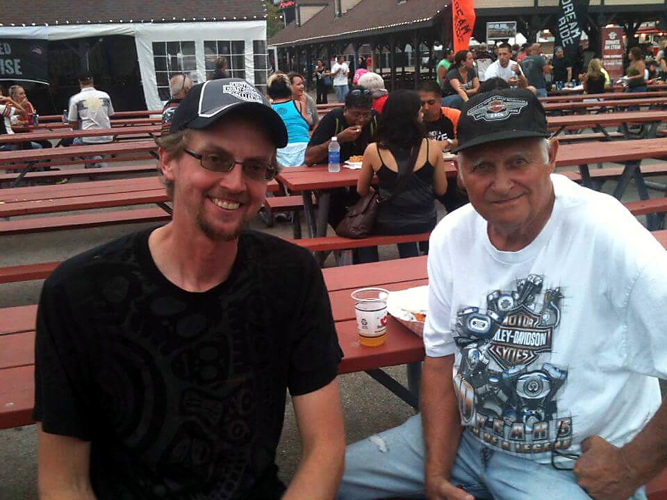 Two men sit at a long red picnic table at an outdoor festival, smiling at the camera.