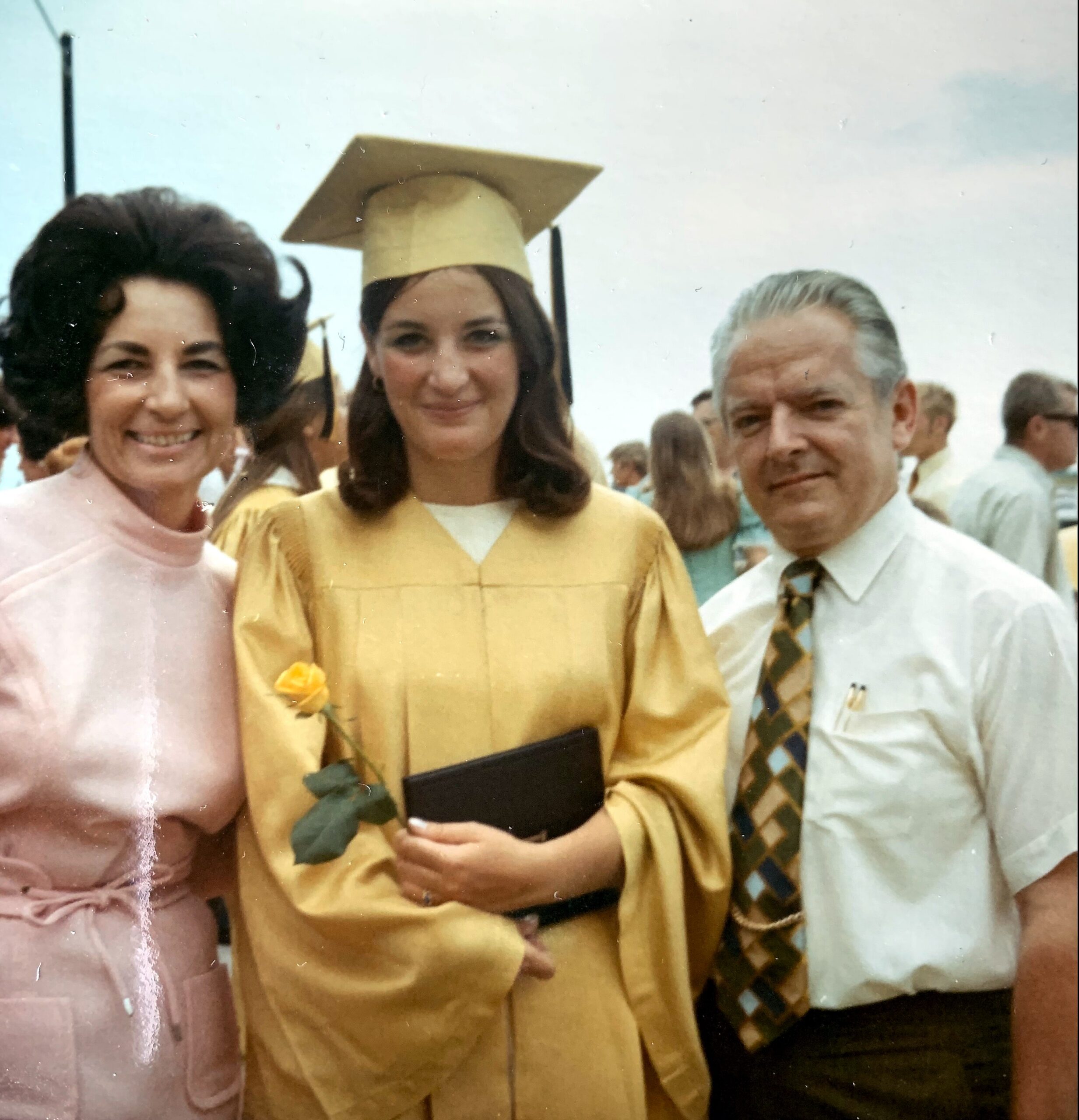 Graduate in a yellow cap-and-gown holds a black diploma folder, flanked by a smiling woman in pink and a man in a light shirt outdoors at a graduation.