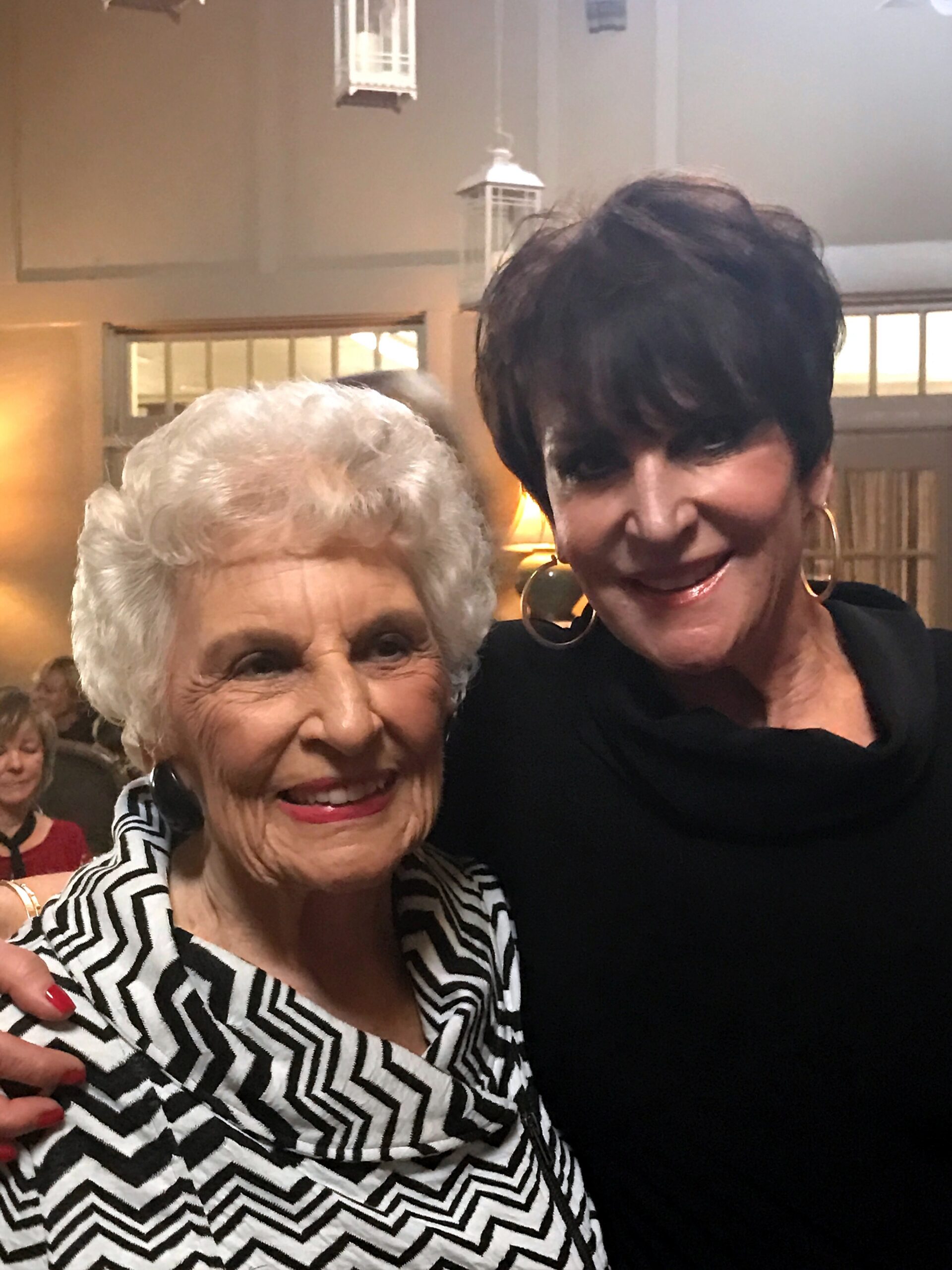 Two women smiling for a selfie at an indoor gathering; one with white hair in a zigzag black‑and‑white blouse, the other with short dark hair in black, large hoop earrings.