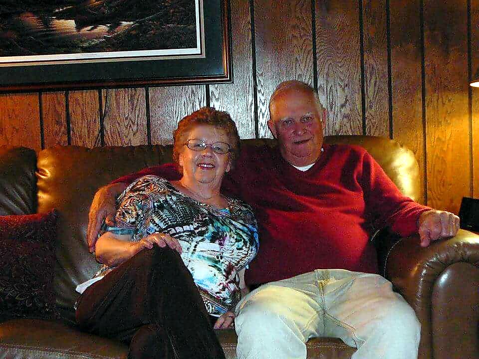 Older couple sitting close together on a brown leather couch in a wood-paneled living room.