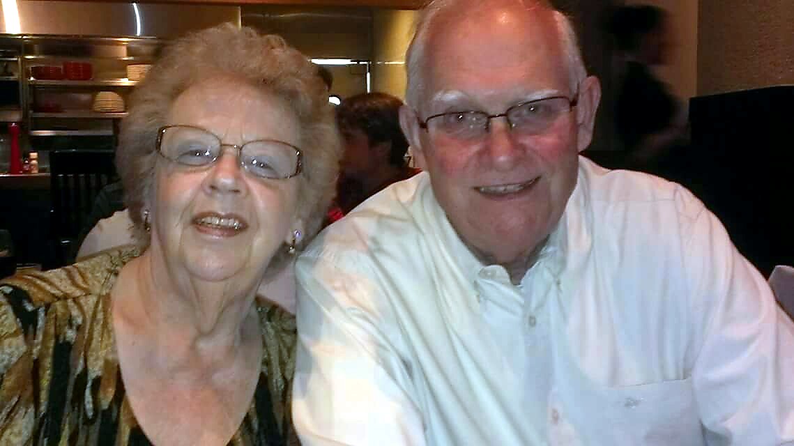 Smiling elderly couple seated at a restaurant, woman with patterned blouse and glasses, man in a white shirt.