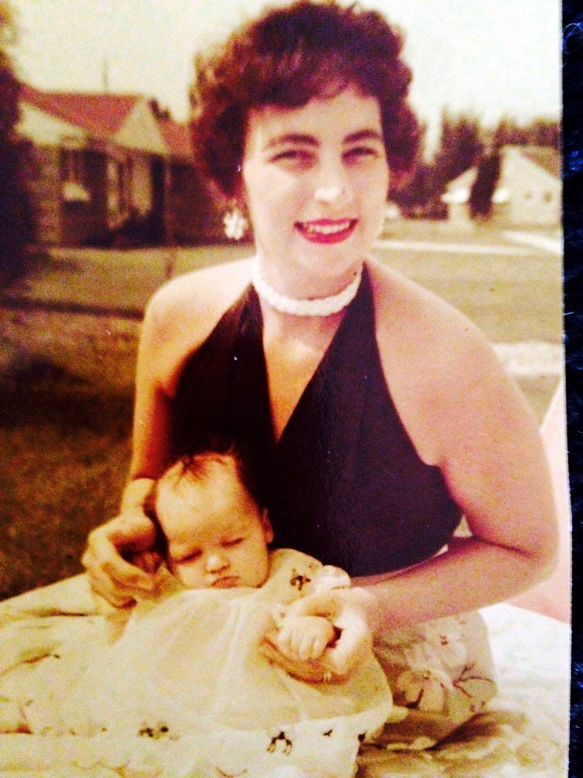 Smiling woman in a dark sleeveless dress and pearl necklace holding a sleeping baby outdoors.