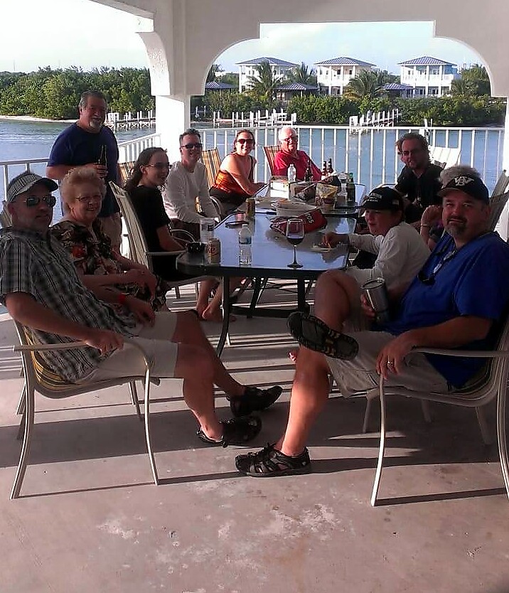 Group of ten adults around a glass table on a covered waterfront deck, sunlit and smiling.