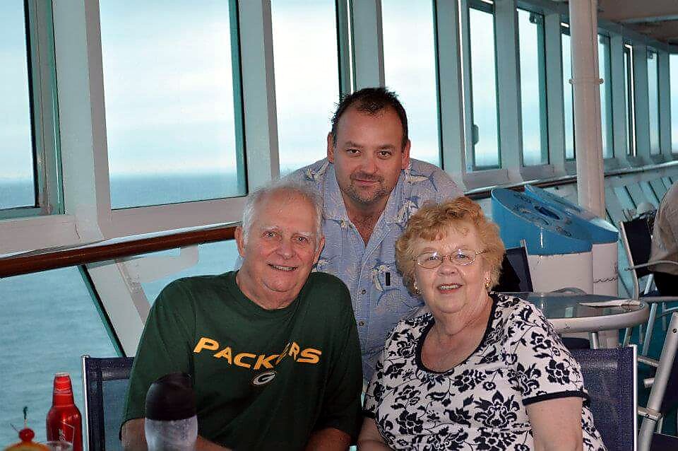 Three adults pose for a photo on a cruise ship balcony, with ocean seen through windows; man in a green Packers shirt sits beside a smiling older woman in a floral top, while another man stands behind them.