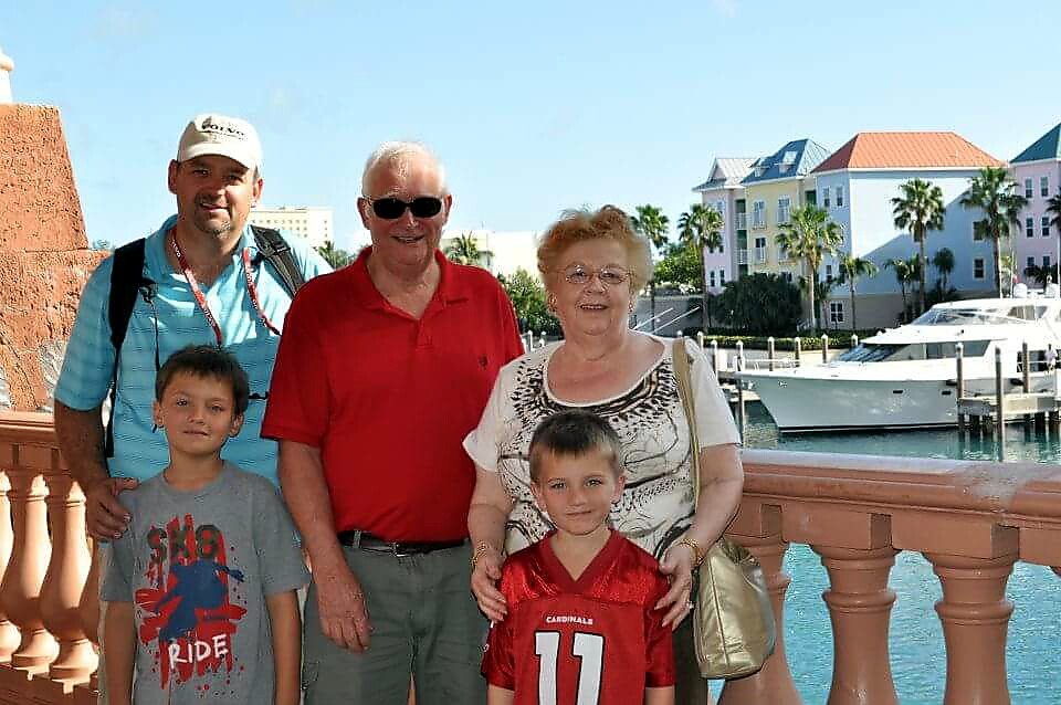 Family of five posing on a sunny waterfront boardwalk with boats and palm trees in the background.