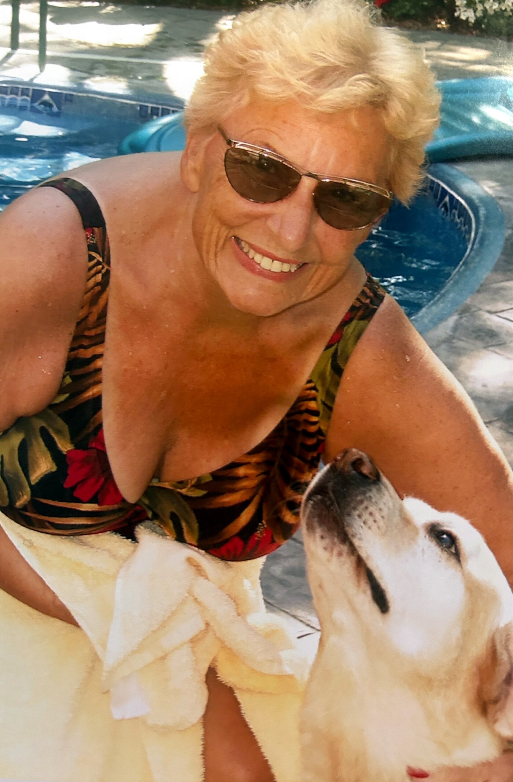 Smiling older woman with sunglasses by a pool; a white dog in a towel sits in the foreground near her.