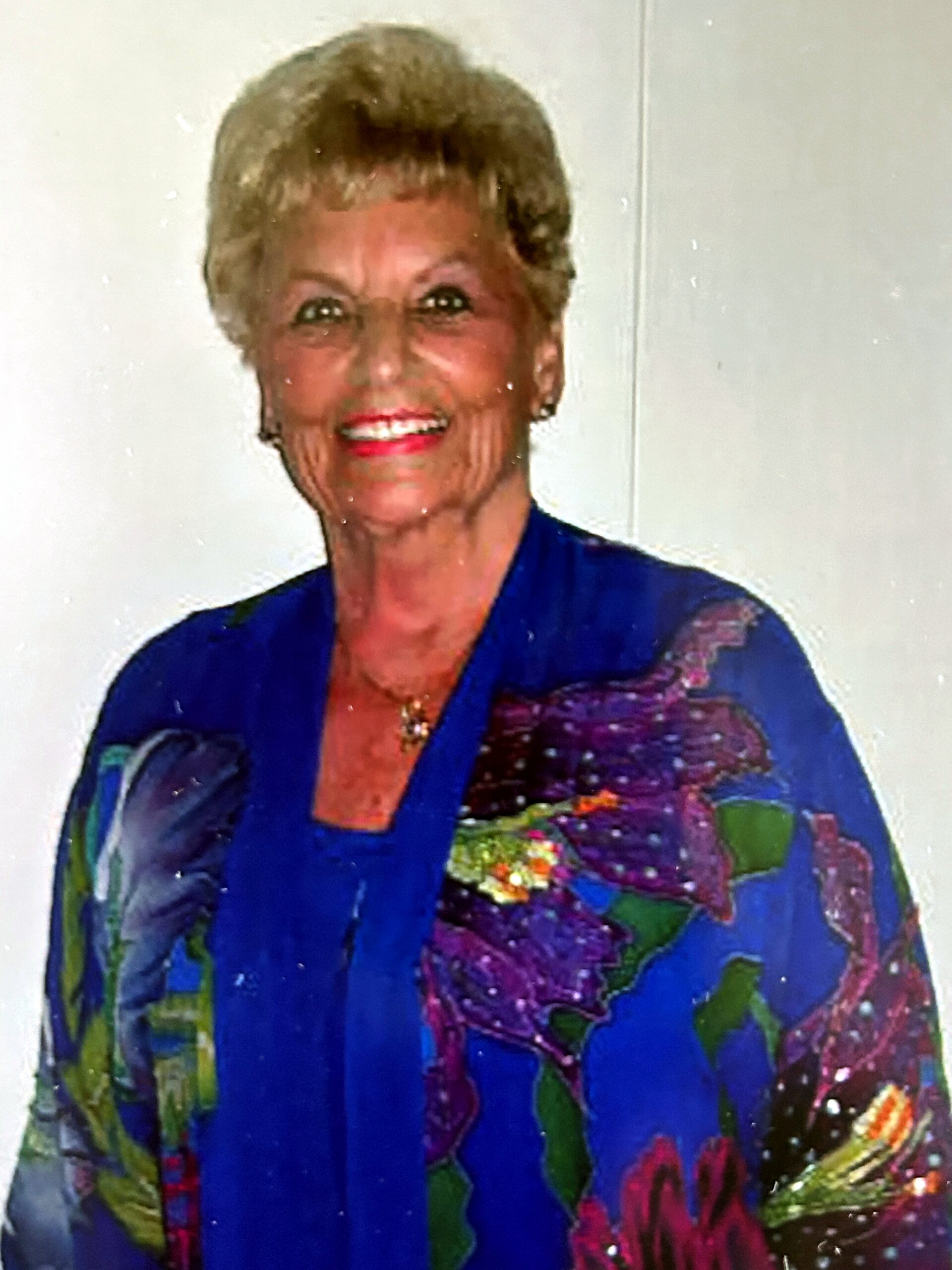 Smiling elderly woman with short blond hair wearing a blue blouse with colorful floral pattern.