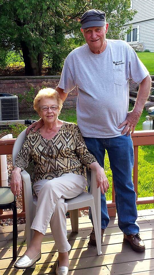 Older woman seated on a patio chair on a wooden deck; man standing beside her with a hand on her shoulder.