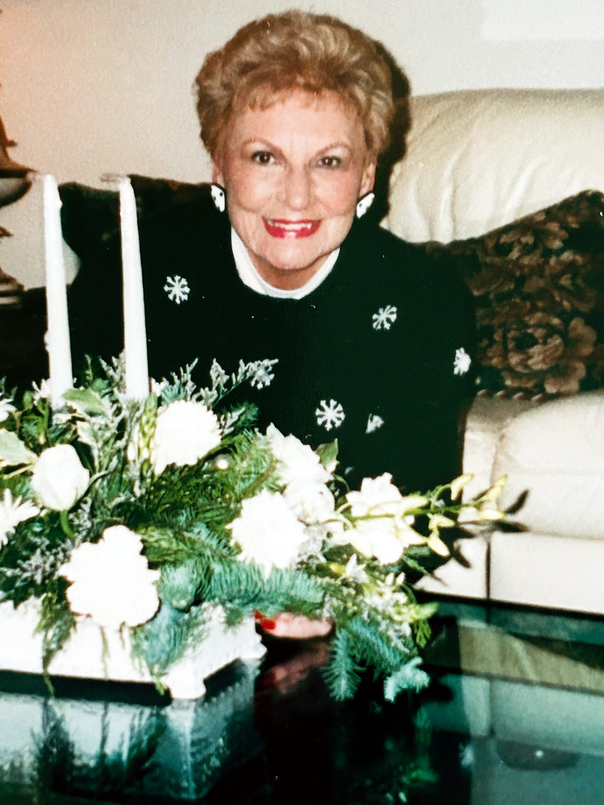 Smiling older woman in a black outfit with snowflake pins, seated behind a white flower centerpiece and candles, festive setting.