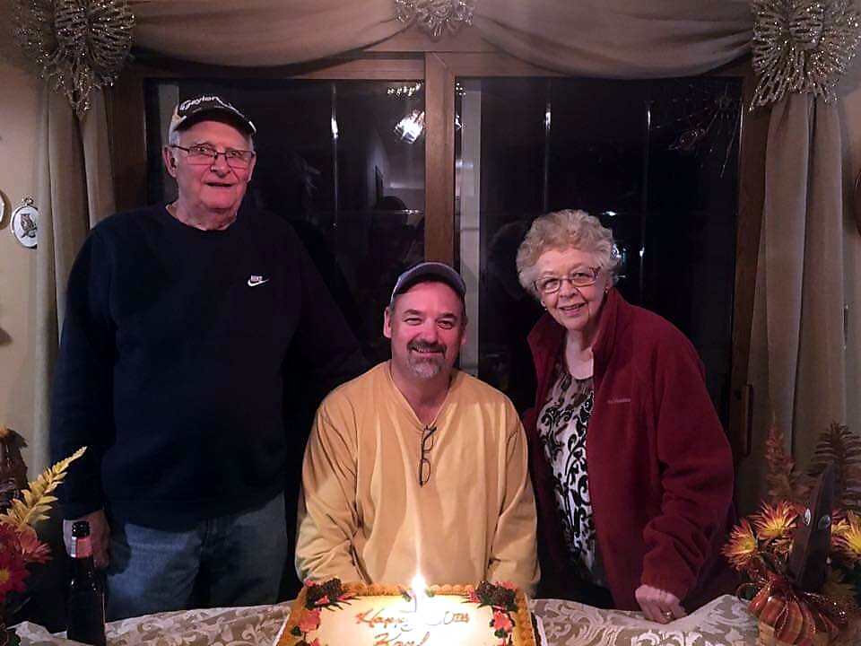 Three adults stand around a birthday cake with one lit candle, indoors by a window.