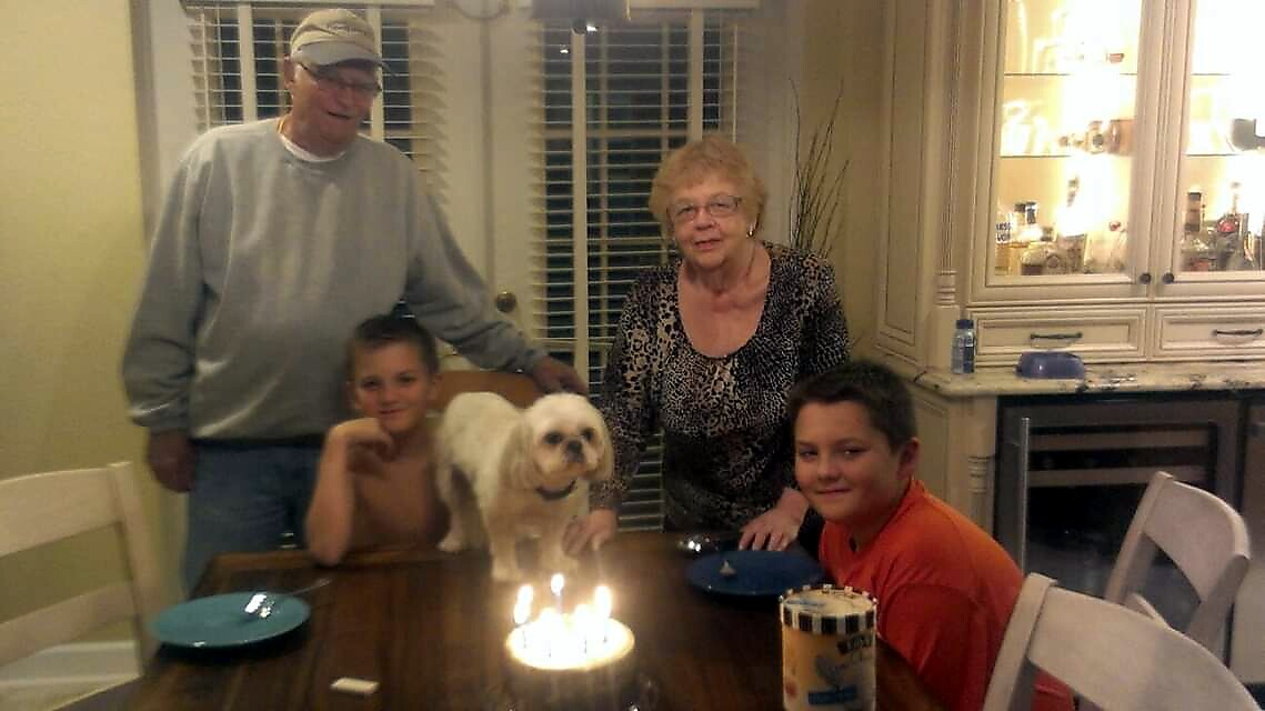 A family gathers around a dining table for a birthday, with a small white dog on the table and lit candles on a cake in front of them.