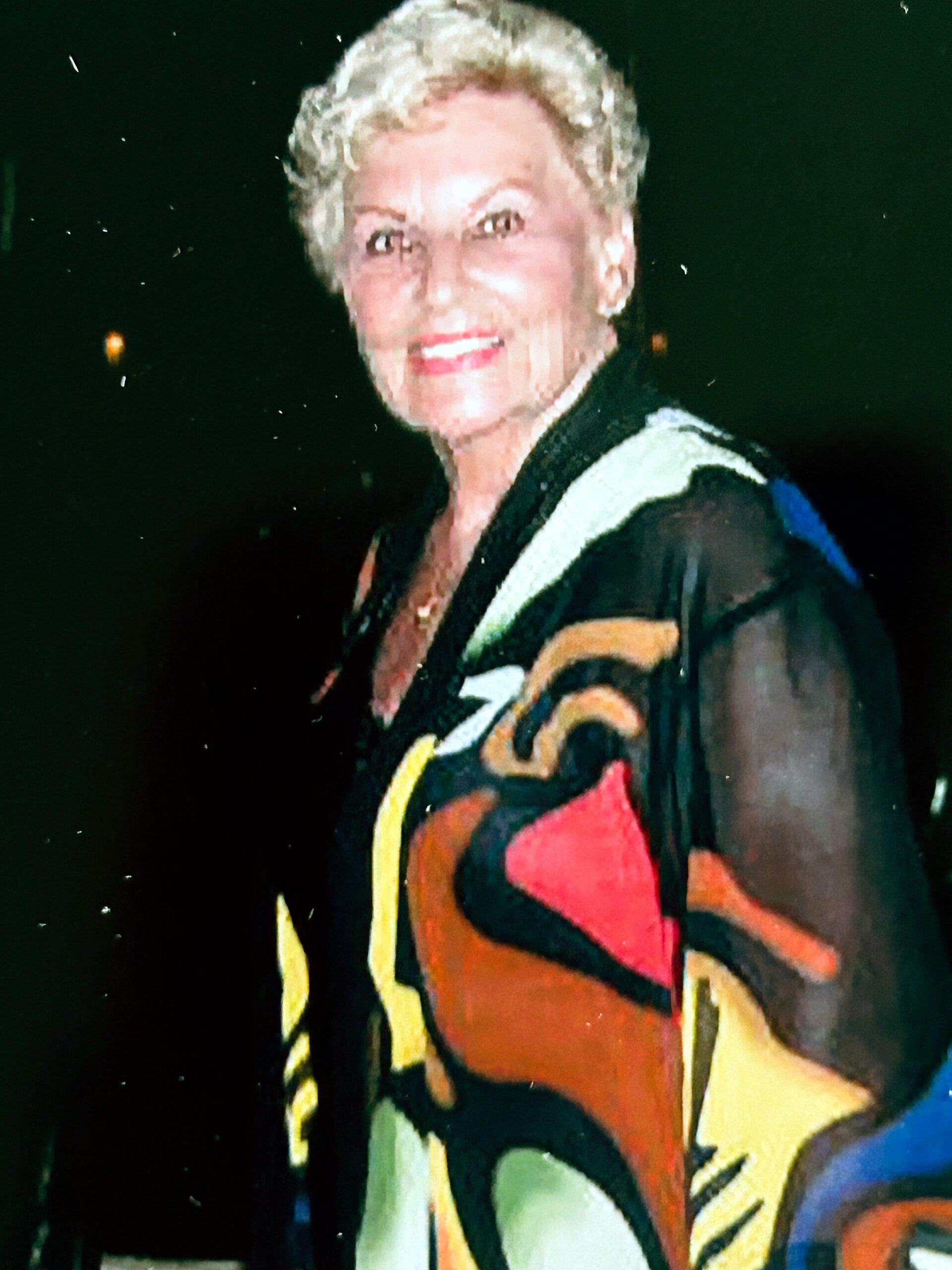 Smiling woman with short blonde hair wearing a bright, multicolored jacket against a dark background.