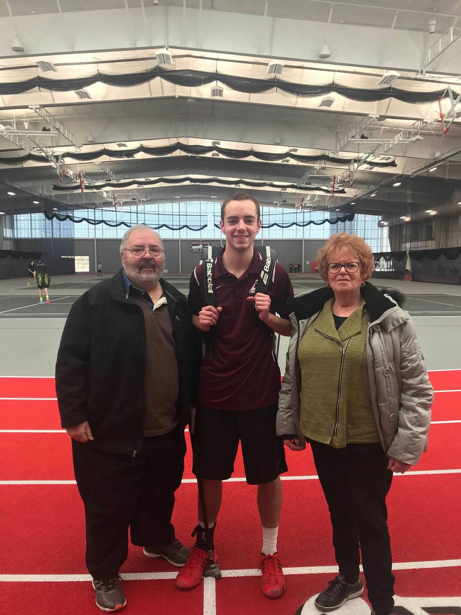 Three people posing on a red indoor sports court: an older man on the left, a smiling young man center in maroon with a backpack, and an older woman on the right.