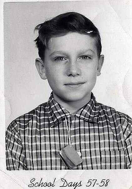 Black-and-white school portrait of a young boy in a plaid shirt with a small tag hanging at his chest, smiling softly, labeled 'School Days 57-58'.