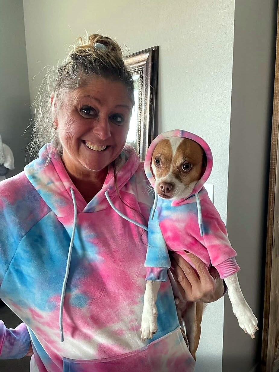 Smiling woman in a pink-blue tie-dye hoodie holding a small dog in a matching hoodie inside a home. Gently grinning at the camera.