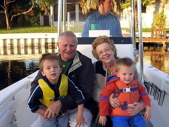 Smiling grandparents with two young boys wearing life jackets sit on a boat at the dock, with a man steering from the cockpit in the background.