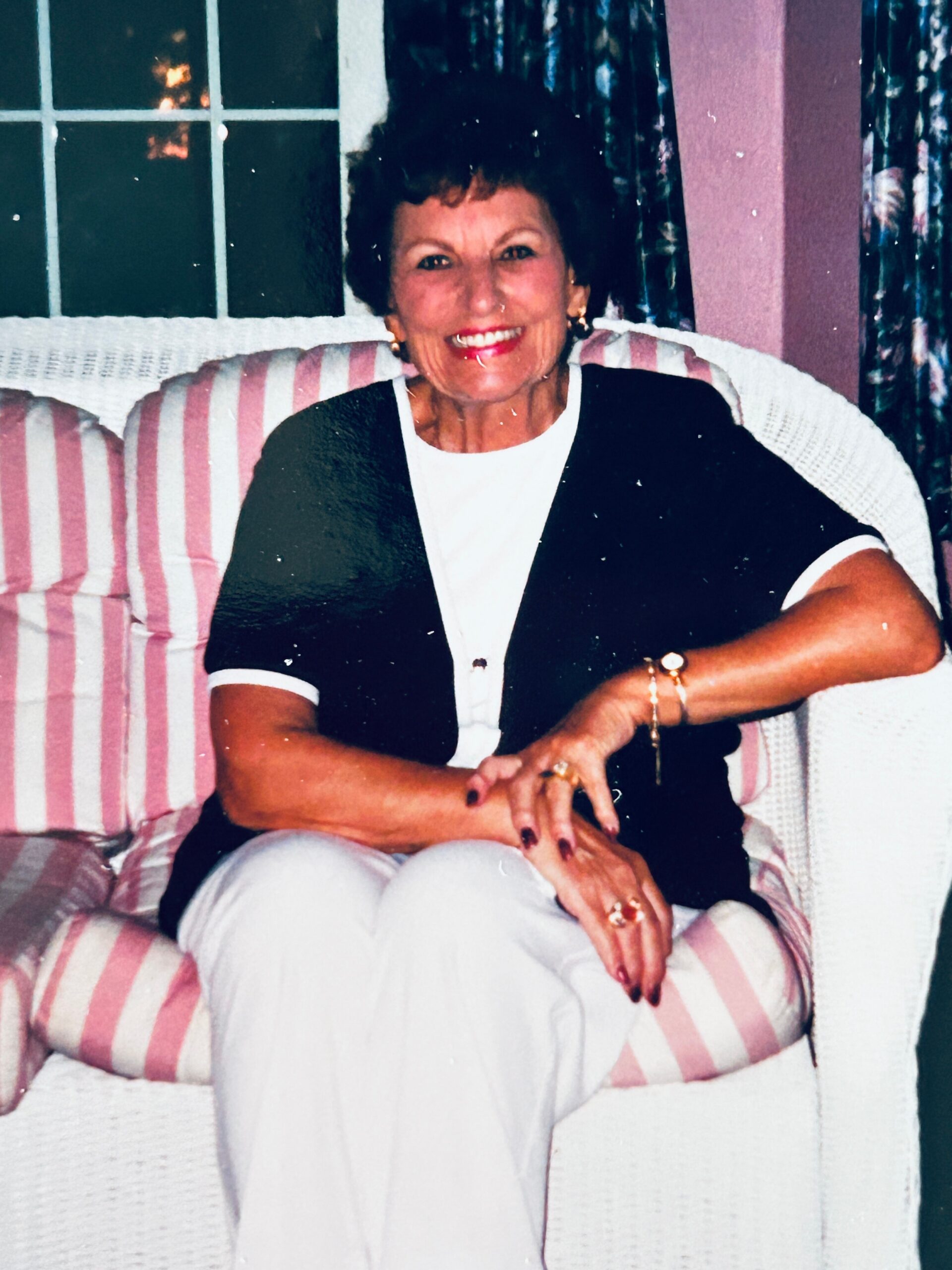 Smiling woman with short dark hair seated on a striped pink outdoor chair, wearing a black and white top.