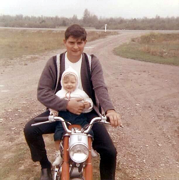 Man wearing a cardigan rides a small motorcycle on a rural dirt road, holding a baby in his lap.
