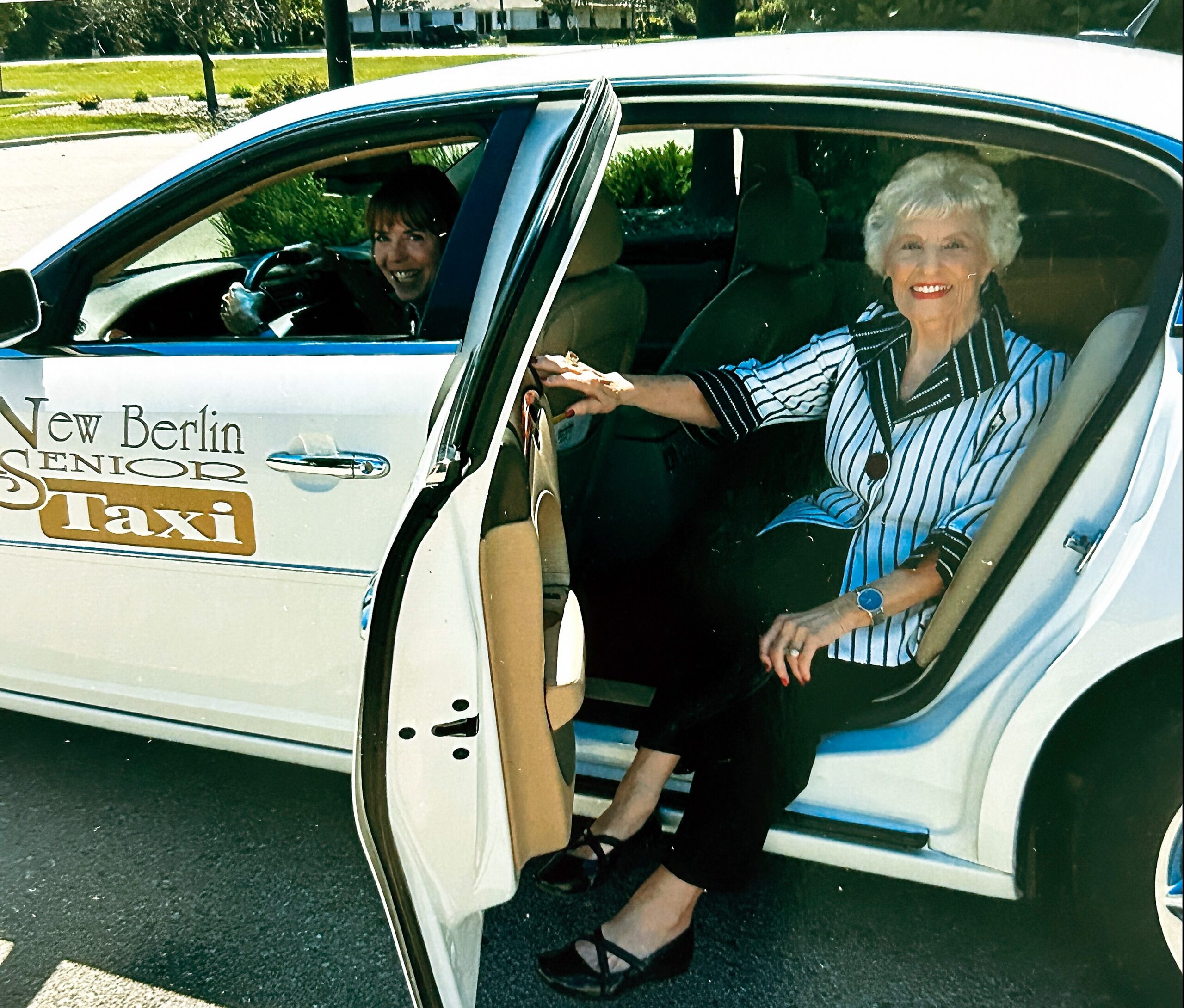 Two smiling senior women sit in a white taxi with its doors open, one driver in the left seat and a passenger in the front, taxi labeled 'New Berlin Senior Taxi' on the side.