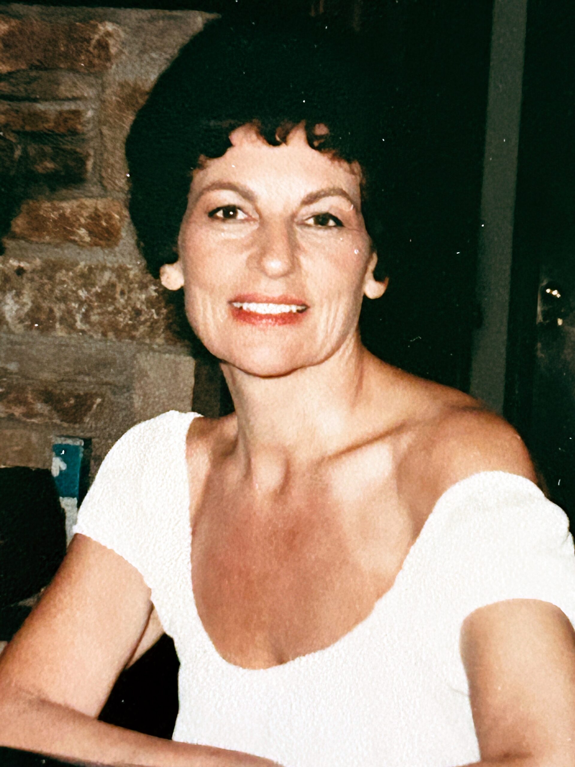 Smiling woman with short dark hair wearing a white off-the-shoulder top, standing indoors near a stone wall.