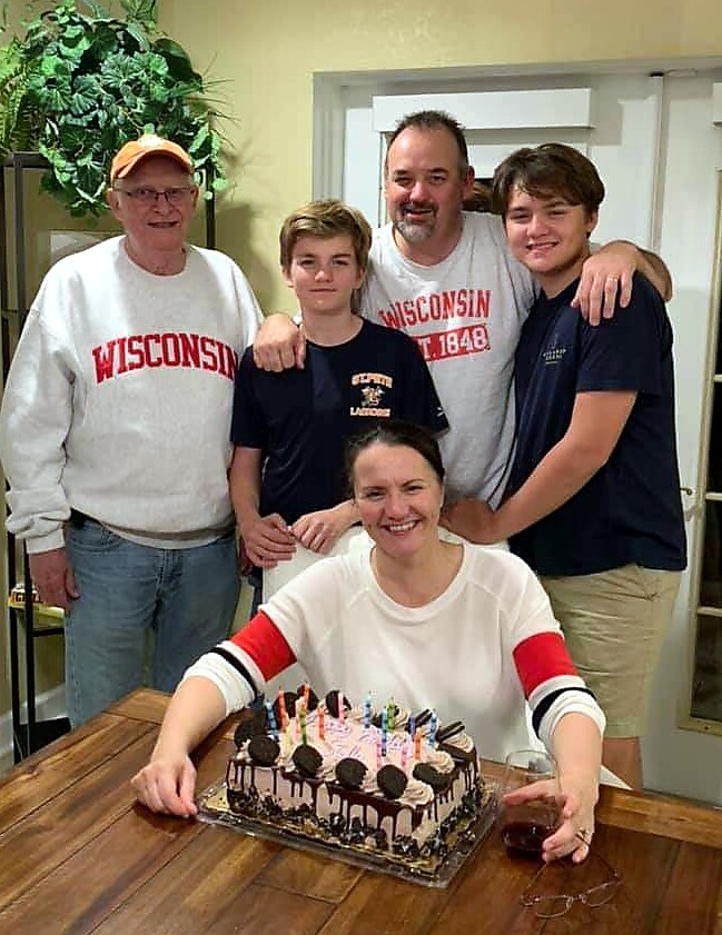 Smiling woman at a wooden table with a chocolate birthday cake and candles, surrounded by four men in Wisconsin shirts.