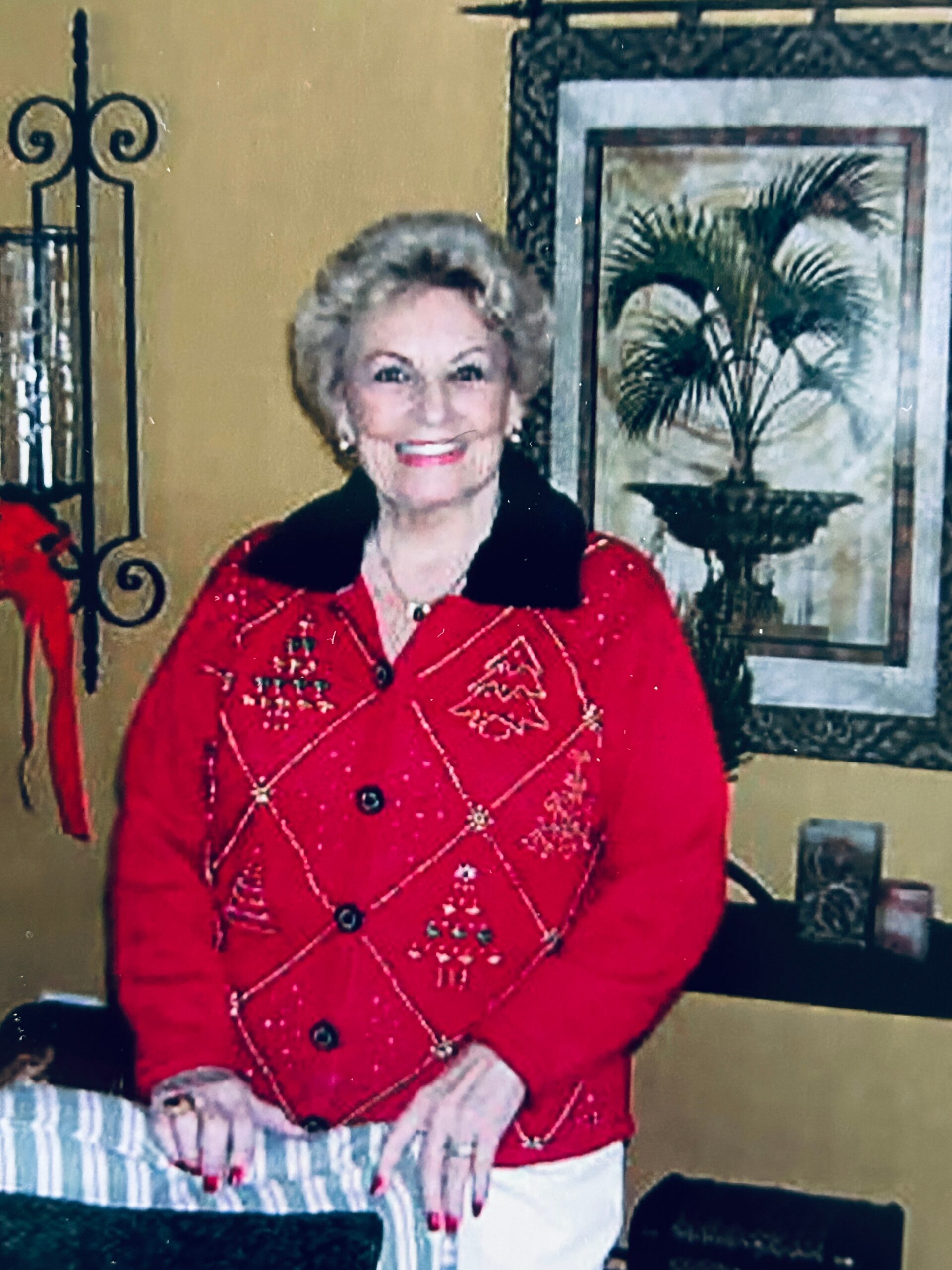 Smiling older woman in a red Christmas sweater posing indoors with a decorative painting behind her.