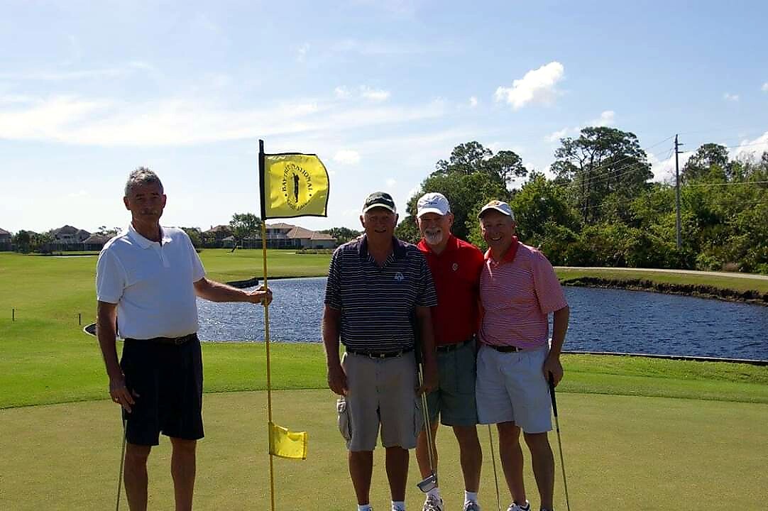 Four men on a golf course posing by a putting green with a water hazard in the background, one holding the yellow flagstick sported in the hole flag.