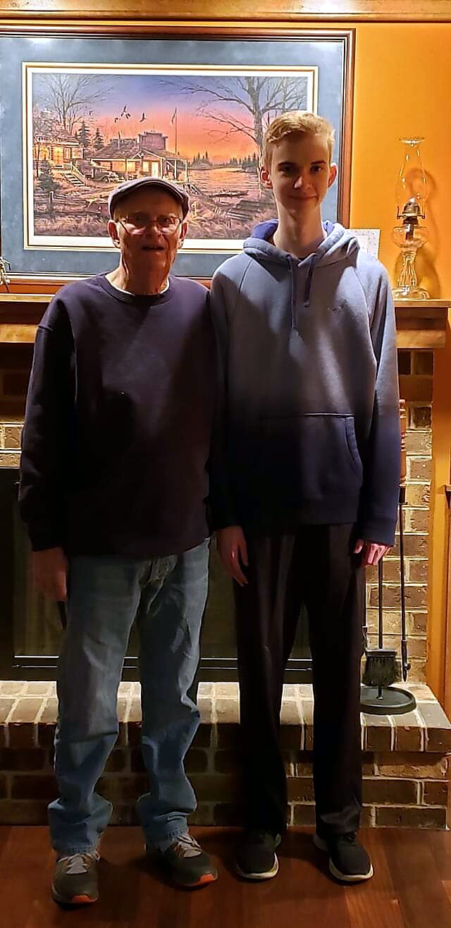 Two men of different ages pose together in a cozy living room, standing by a brick fireplace with a framed landscape painting above it.