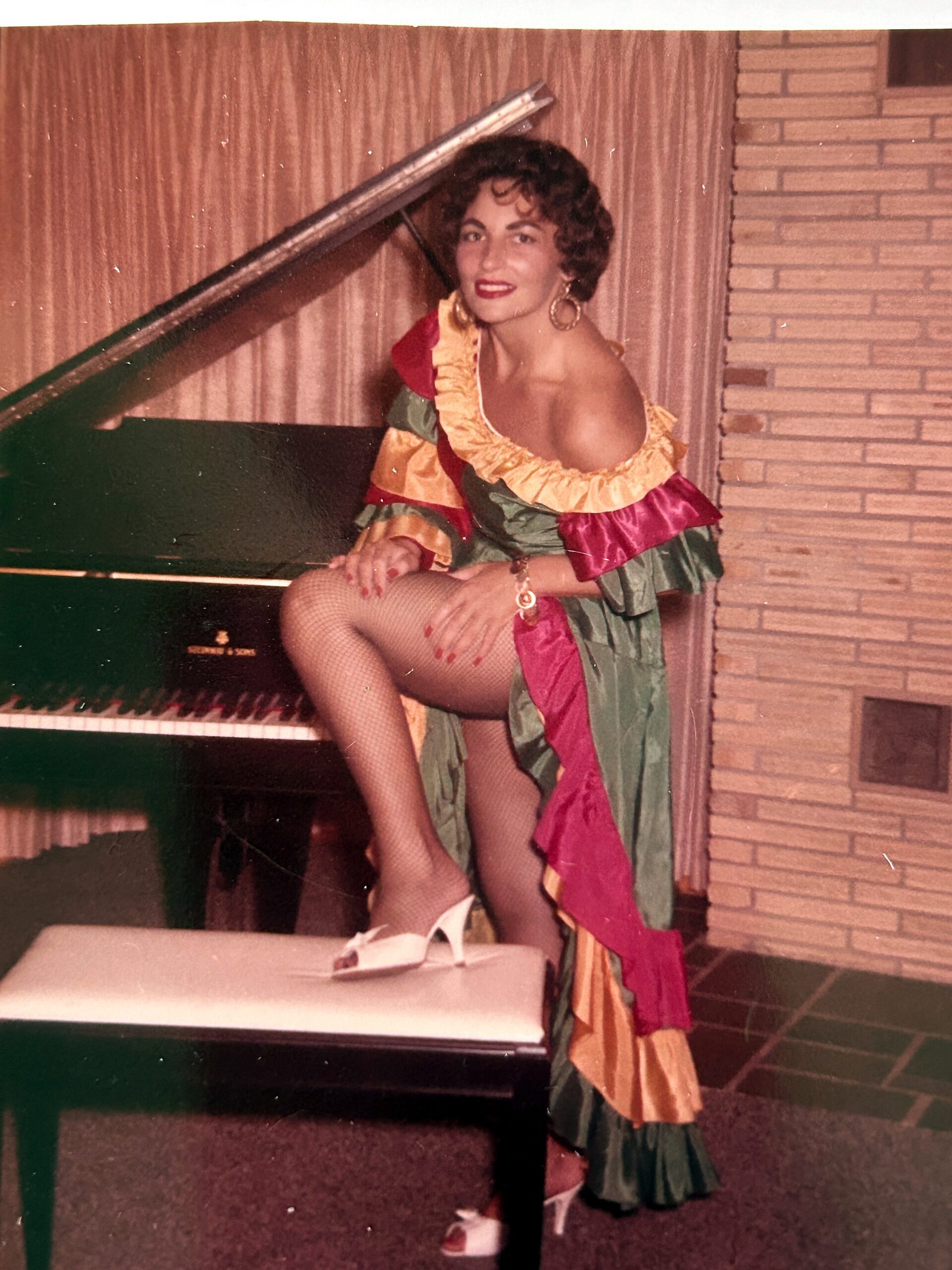 Woman in a colorful ruffled off-shoulder dress with fishnet stockings sits by a grand piano, wearing white heels and hoop earrings.