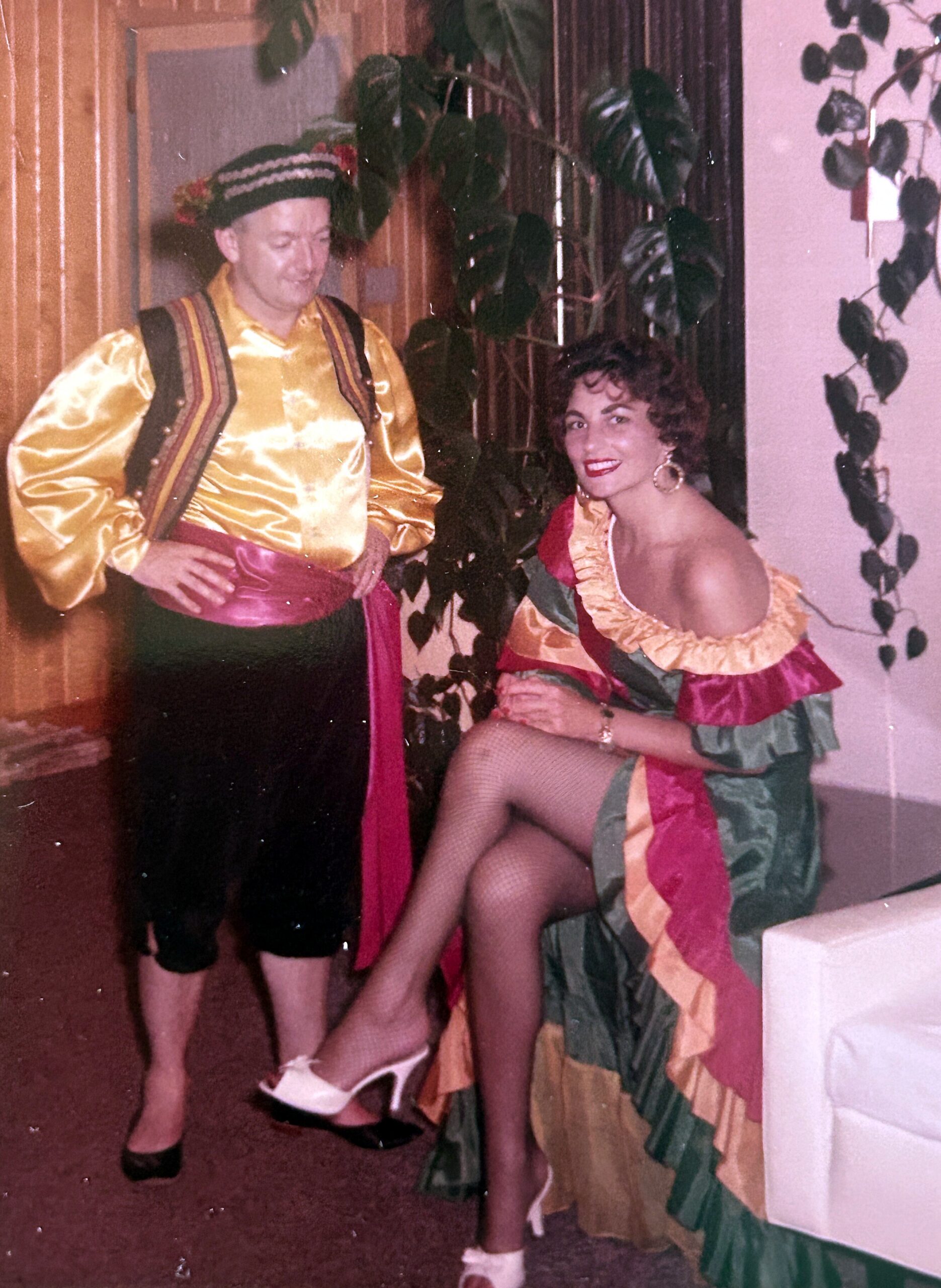 Man in a shiny gold satin shirt with a colorful sash and striped flowered hat stands beside a woman in a bright, multi‑colored ruffled dress at a party.