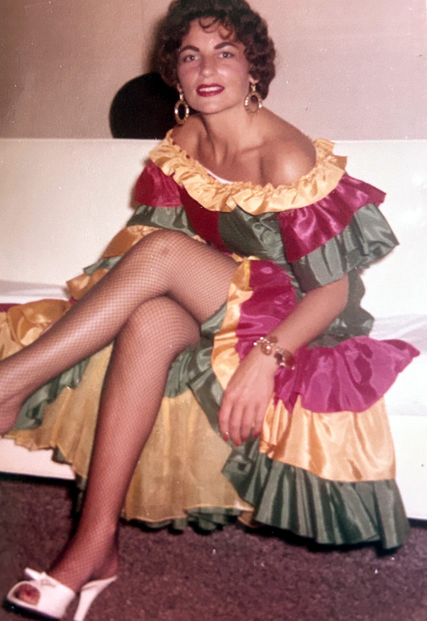 Woman posing in a colorful, ruffled off-the-shoulder dress with fishnet stockings and large hoop earrings, seated on a carpeted floor.
