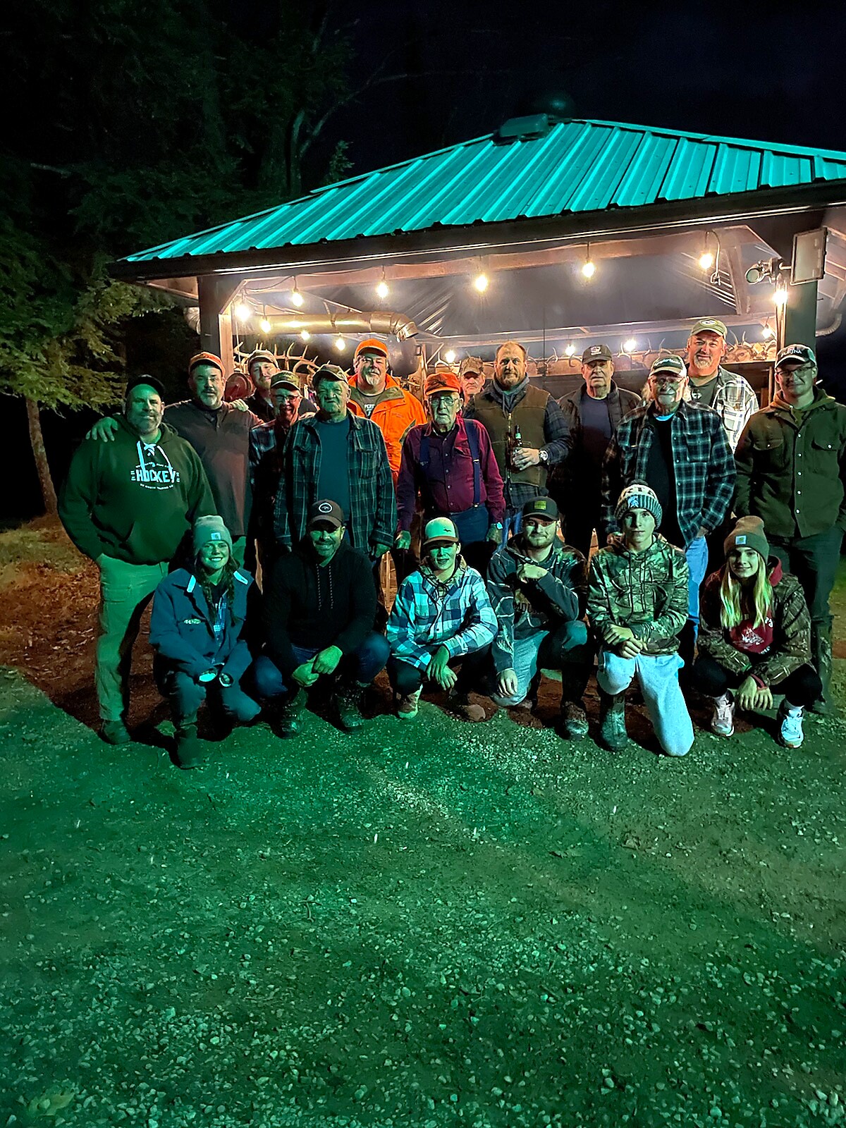 Group of men and women posing outside a lit cabin with string lights at night, wearing outdoor clothing and hats for a gathering or event.
