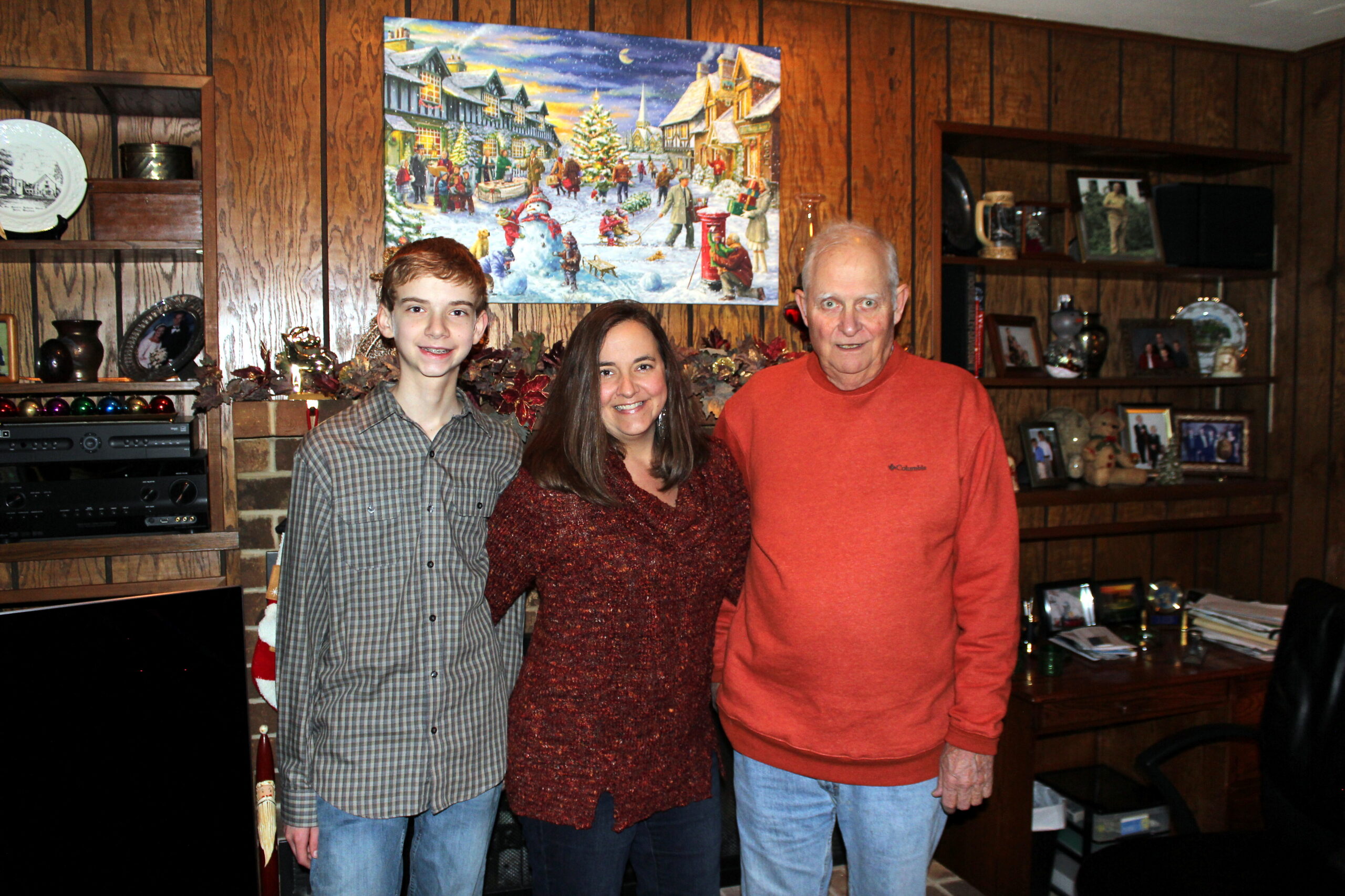 Three people stand together and smile for a photo in a wood-paneled living room with Christmas decor and a large holiday painting behind them.