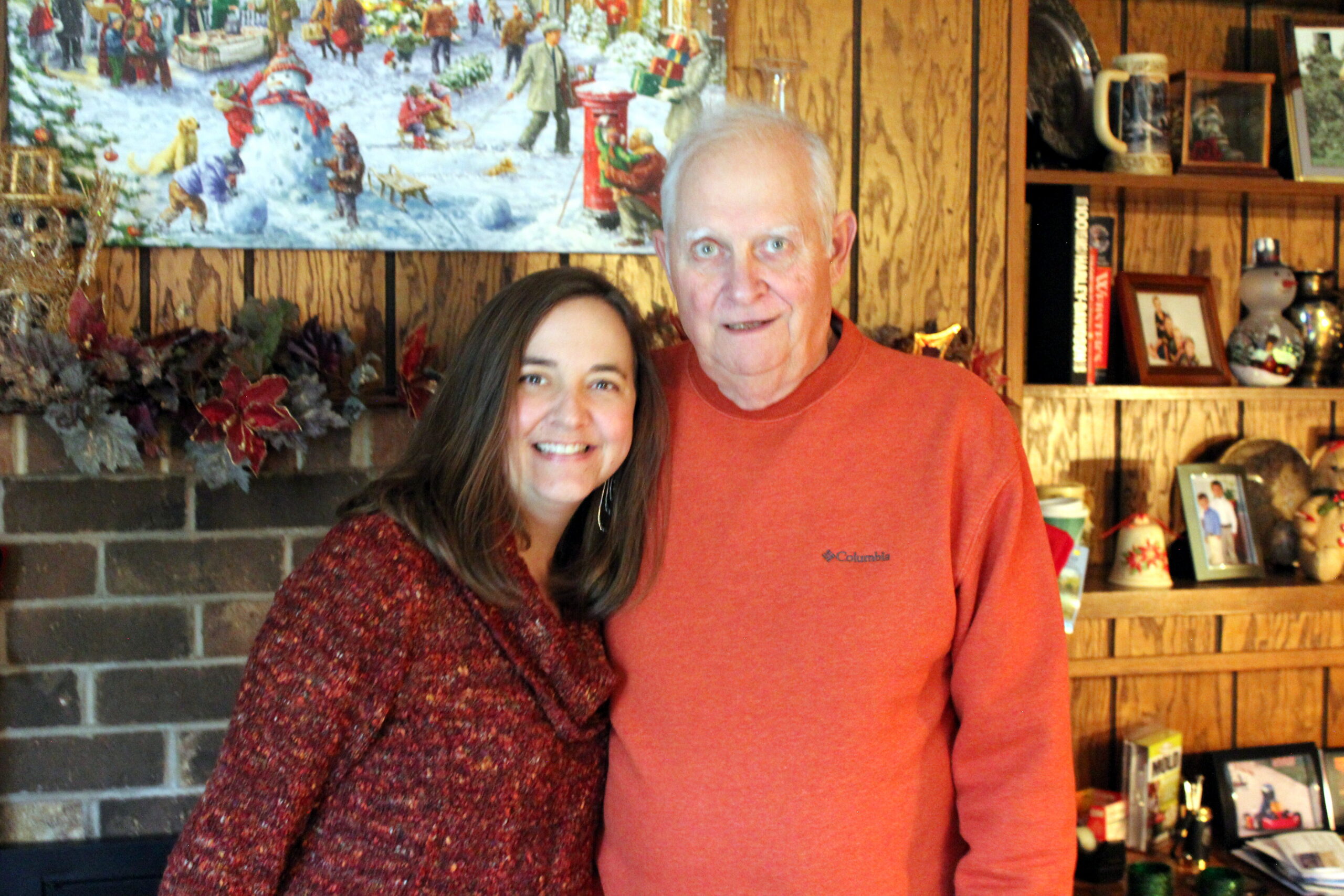 Smiling woman and man stand together in a cozy living room with holiday decor and wooden shelves in the background.