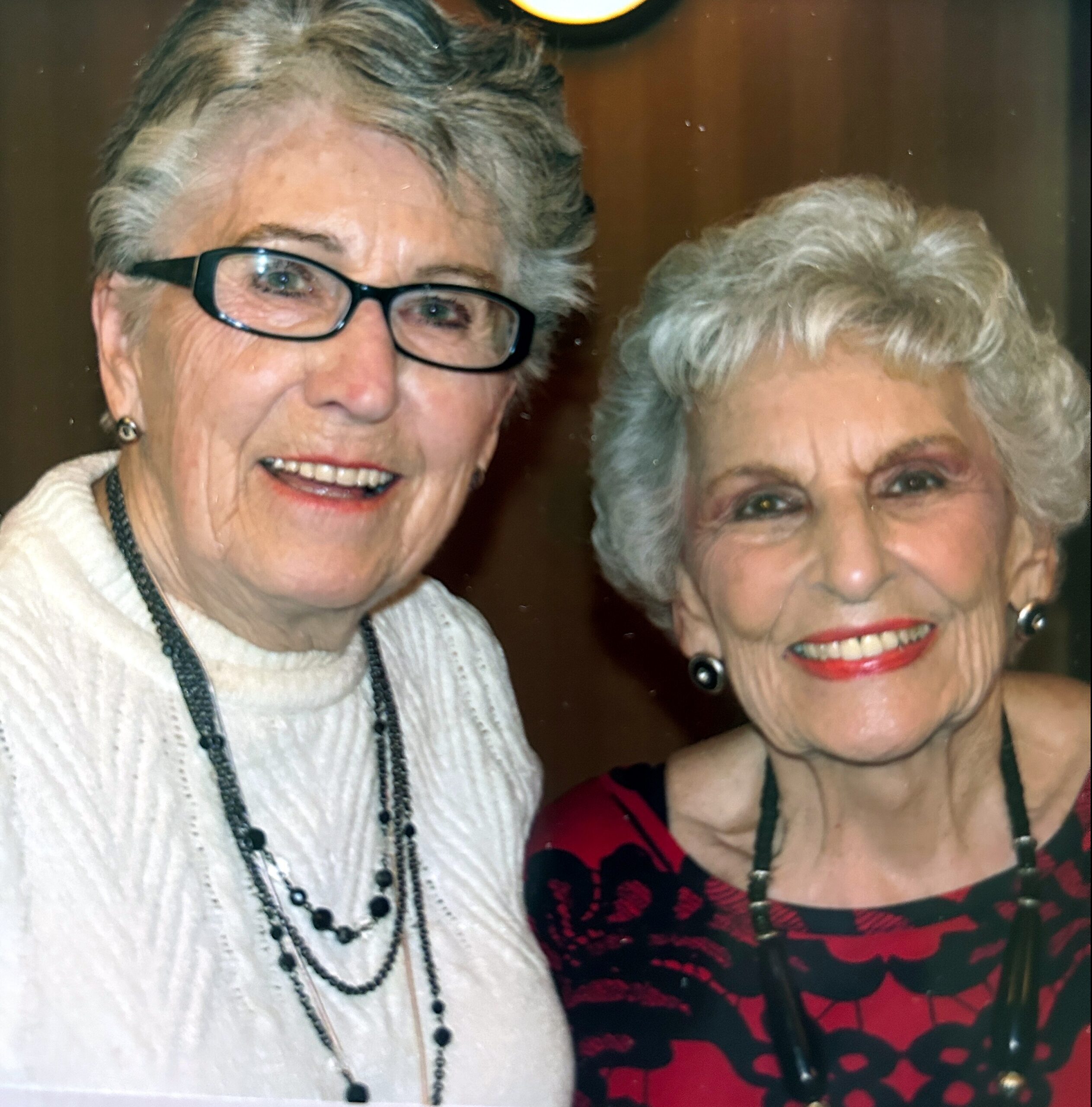 Two elderly women smiling for a close-up selfie, one wearing glasses and a white sweater, the other in a red patterned top with jewelry.