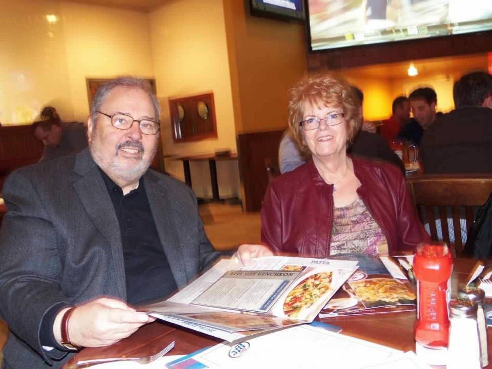 Smiling man in a blazer and woman in a red jacket sit at a restaurant table, looking at menus.