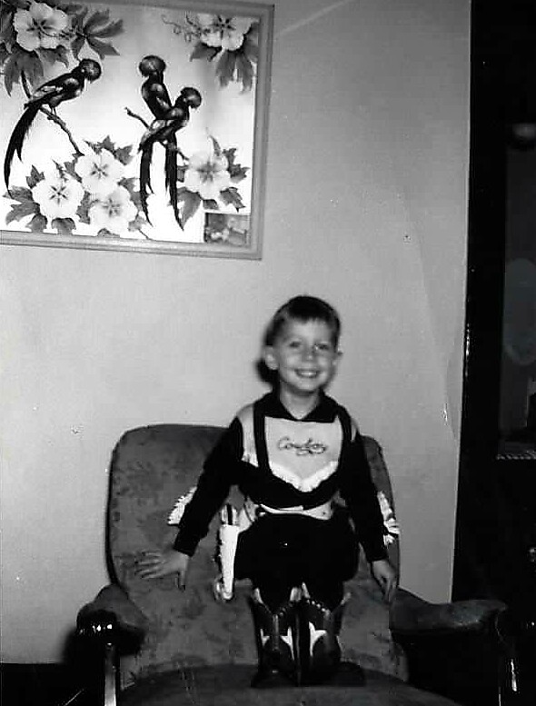 Young boy in a cowboy costume sits on a cushioned armchair, smiling at the camera in a living room.