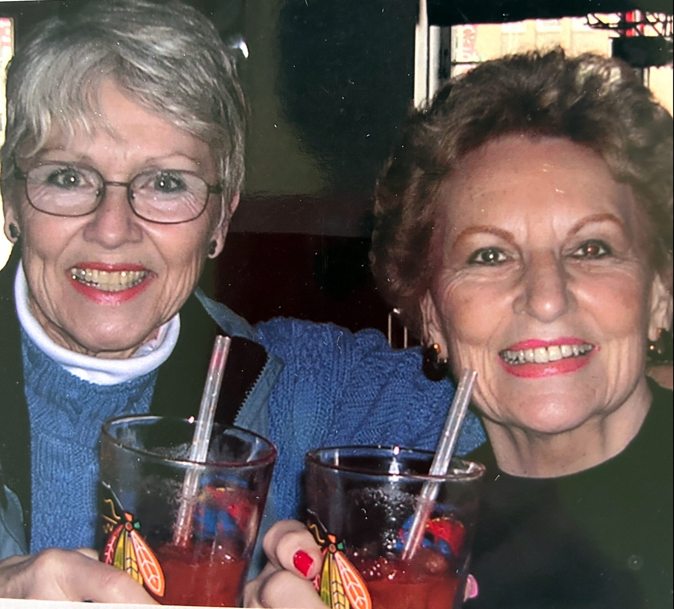 Two smiling elderly women pose for a close-up photo, wearing blue sweaters and holding colorful party poppers with orange decorations.