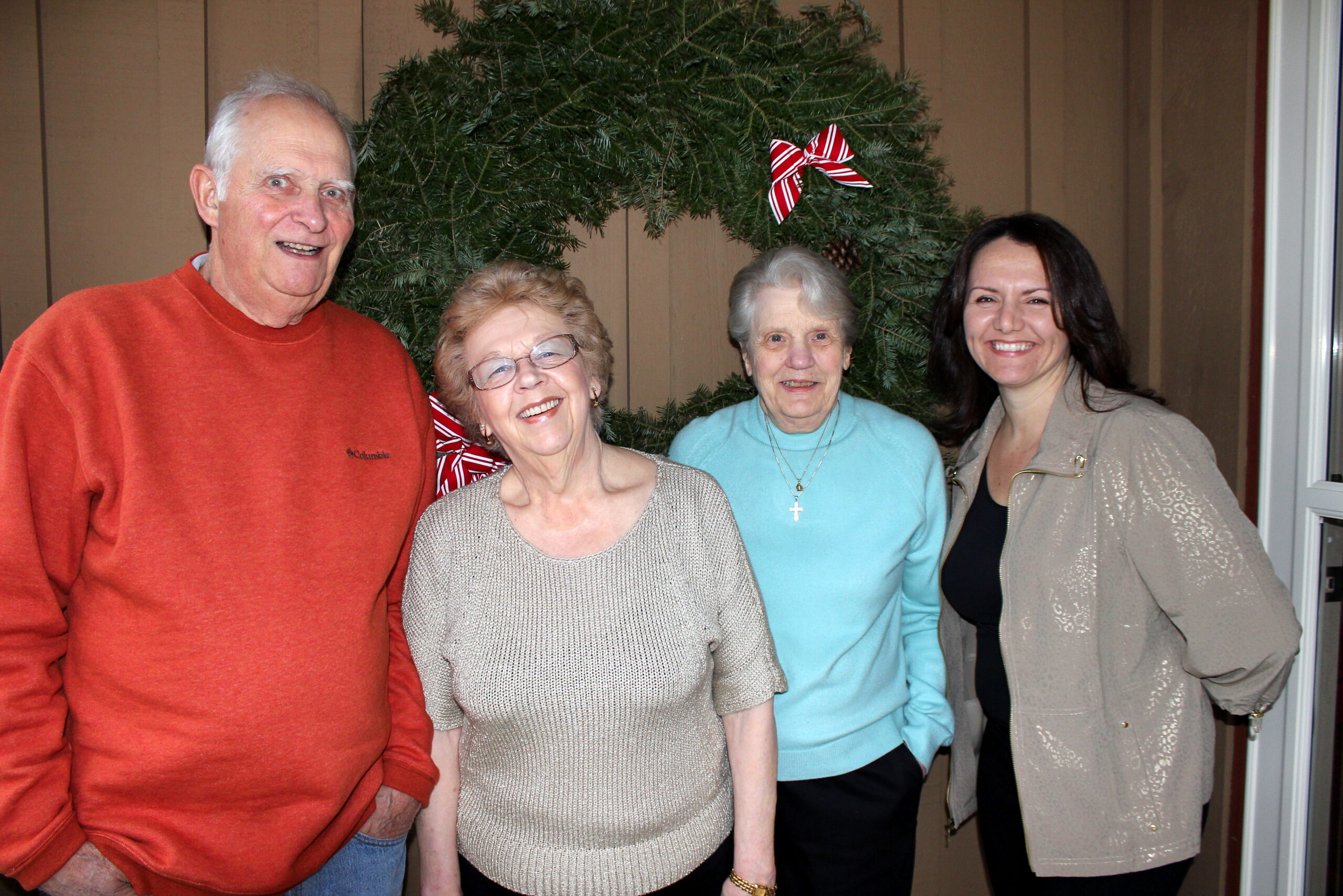 Four adults smiling in front of a large evergreen Christmas wreath with a red-and-white striped bow.