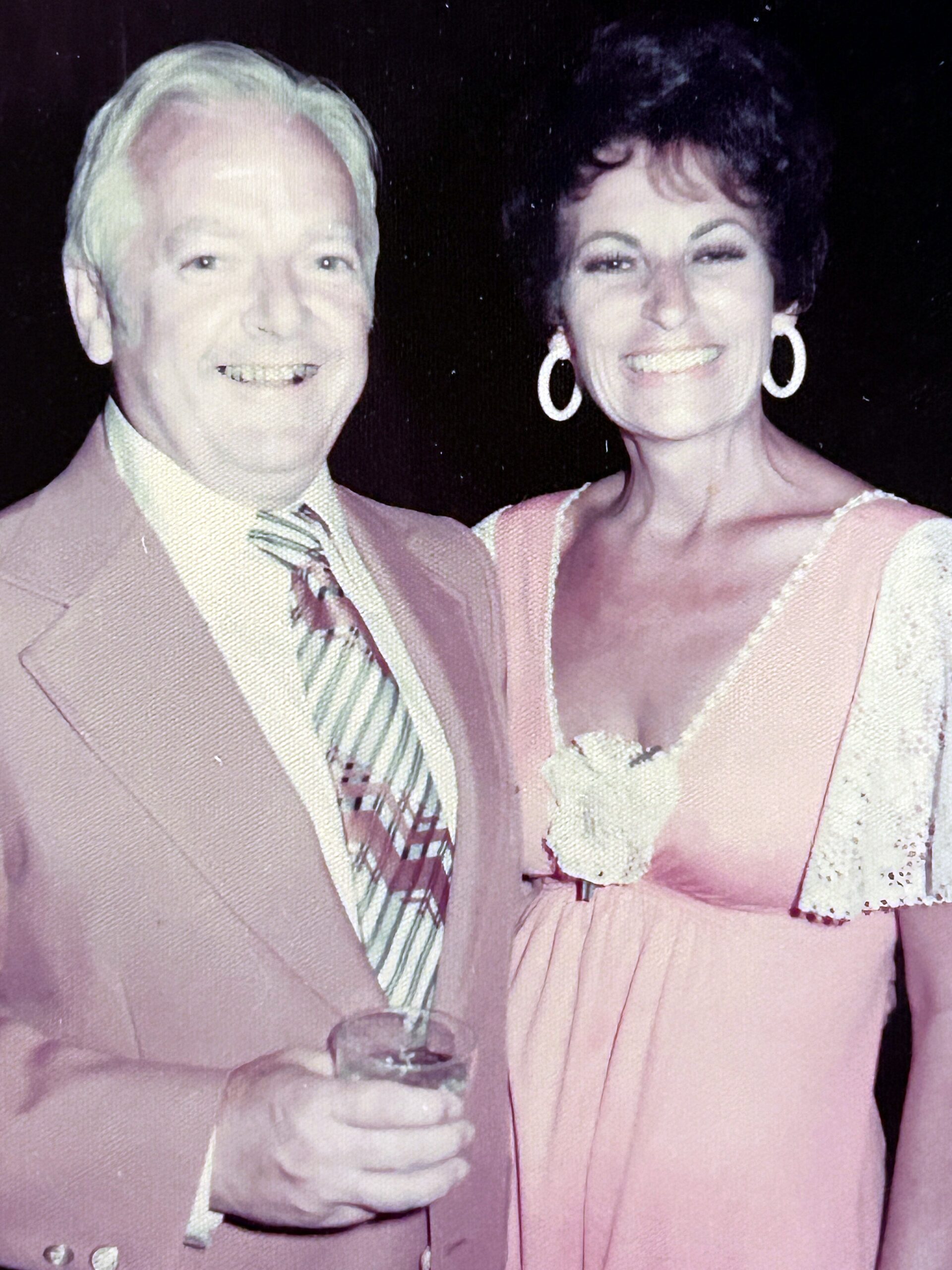 Smiling older couple in pink formal attire posing at a celebration, the man in a suit and tie and the woman in a pink dress holding a champagne flute.