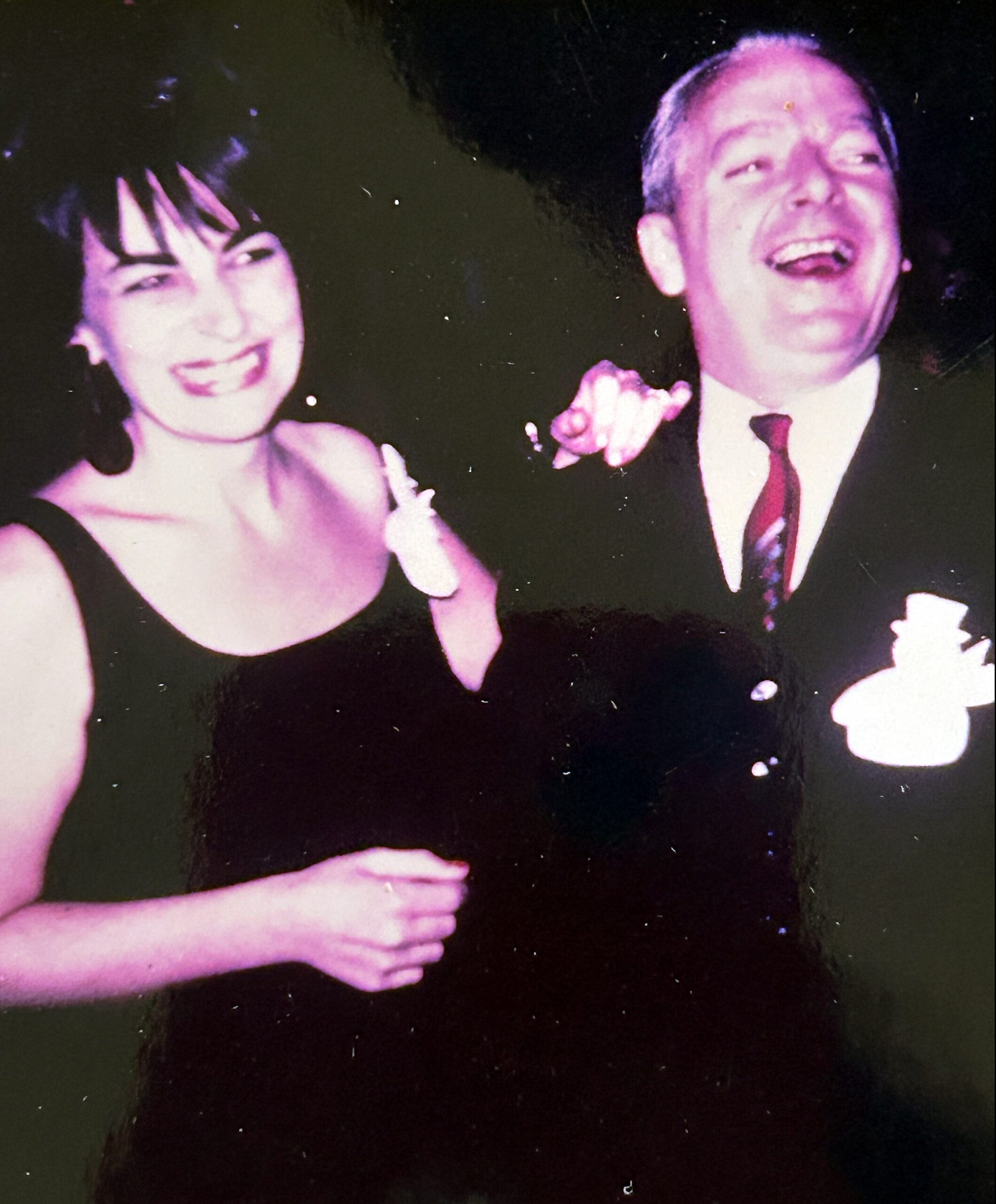 Smiling woman in a dark dress and a man in a suit laughing together at a nighttime event, arms around each other.