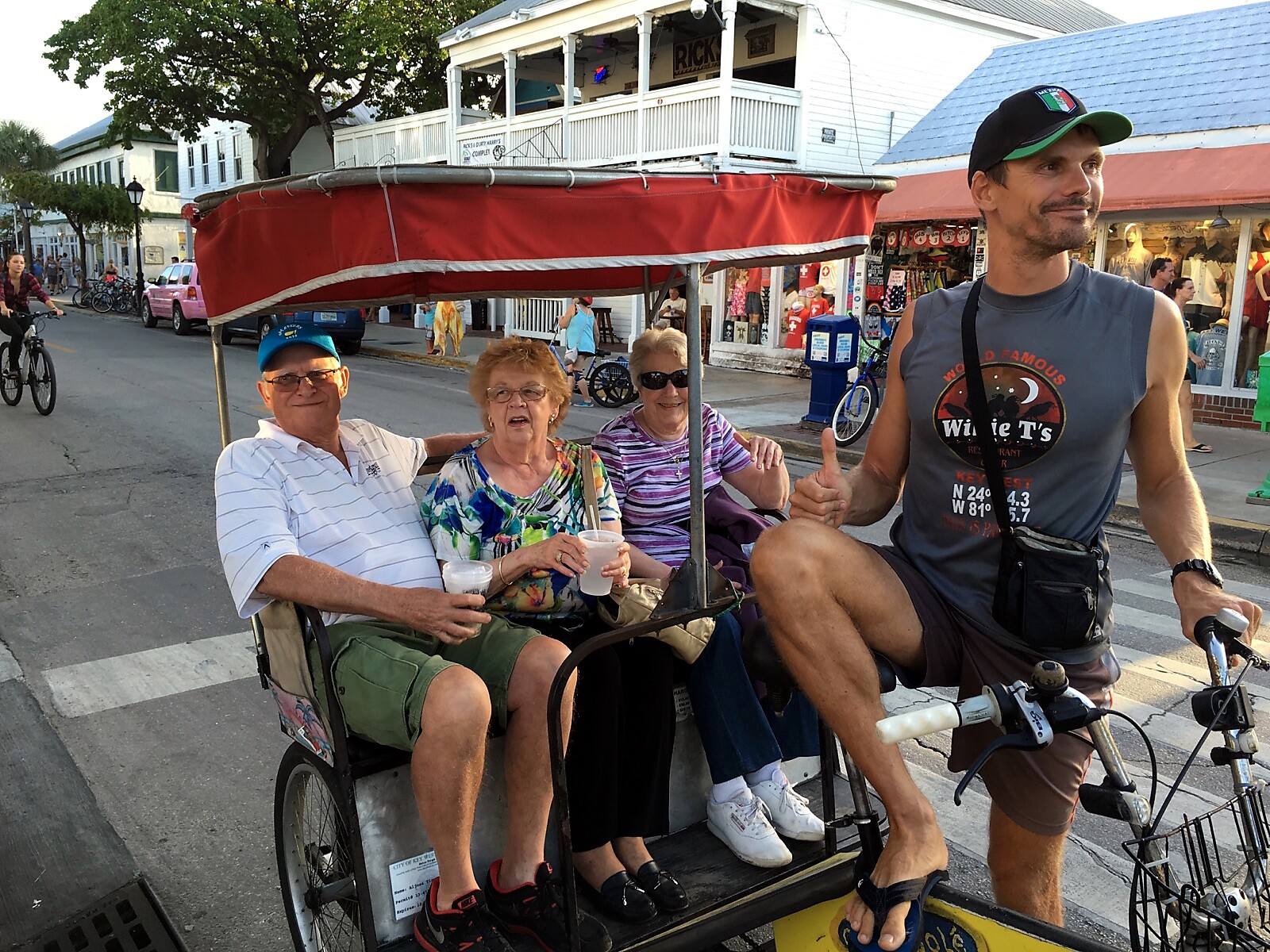 Three seniors ride in a red-canopy pedicab while a man on a bicycle guides them down a sunny street with shops in the background.