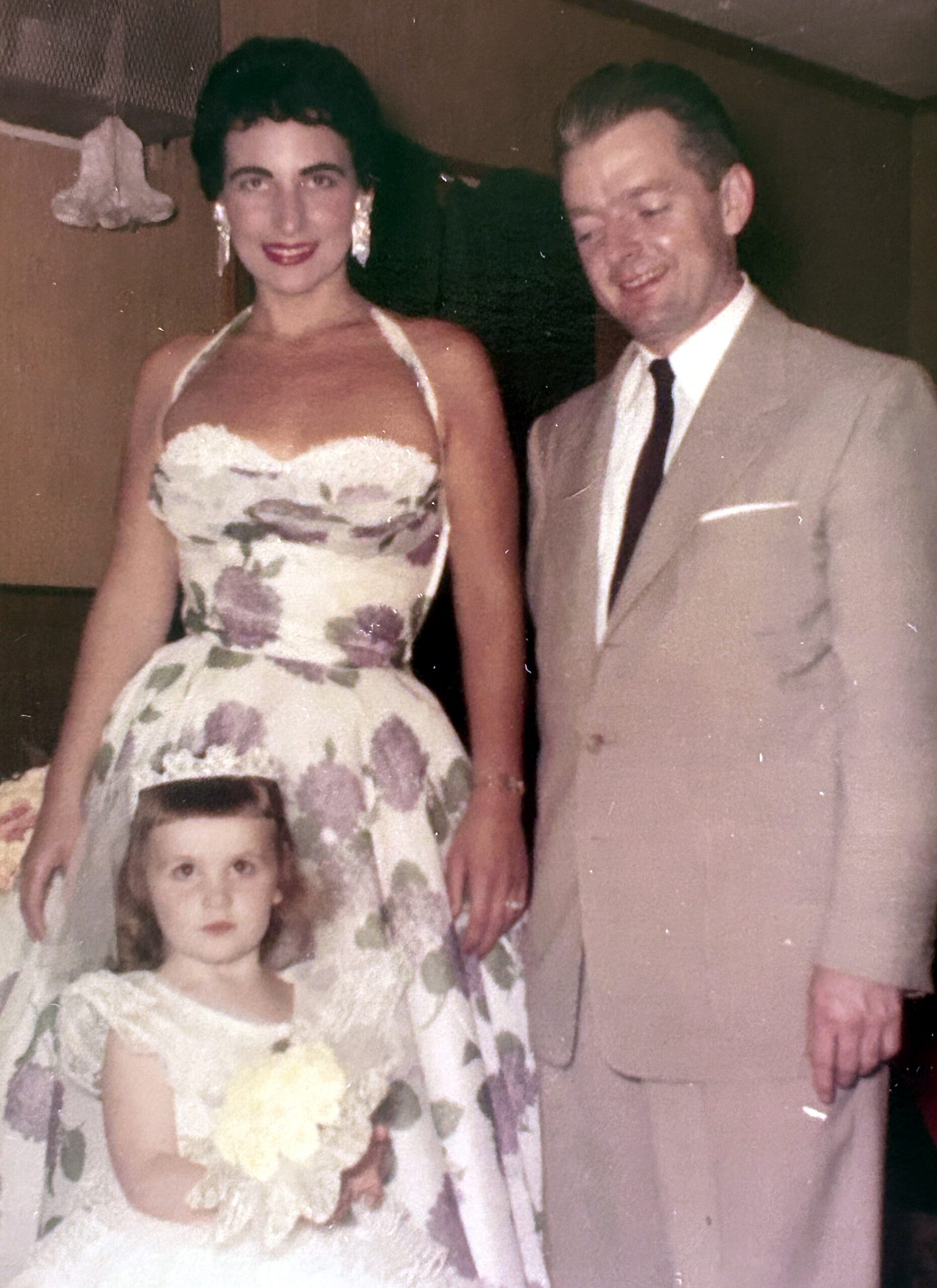 Woman in a floral dress and man in a beige suit pose with a young girl in a white dress indoors at a family event.