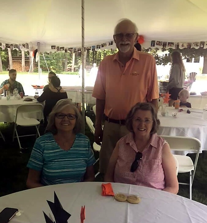 Three seniors under a white tent at an outdoor party: two women seated at a round table, a man standing behind them smiling.